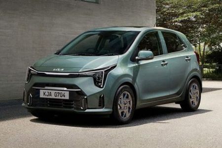 A light green Kia hatchback, with license plate KJA 0704, is parked on a paved area beside a concrete building and greenery, showcasing its modern and compact design.
