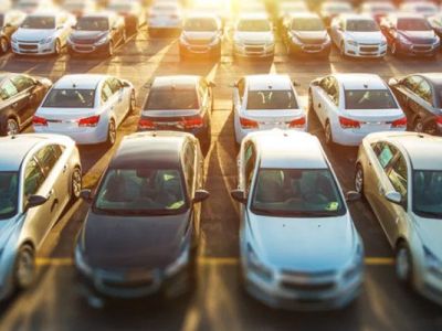 Sunlit parking lot filled with rows of parked cars, featuring a mix of white, black, and silver vehicles.