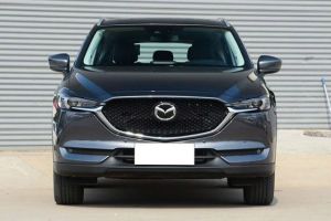 Front view of a dark gray Mazda SUV, parked in front of a corrugated metal wall, featuring its grille, headlights, and sleek front design with a blank license plate.