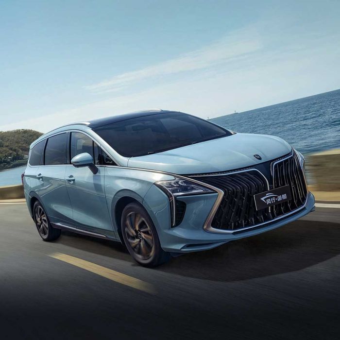 A light blue MPV drives along a coastal road, with the sea and clear sky in the background, showcasing its sleek design and dynamic appeal.