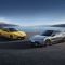Two sports cars, one yellow with gray stripes and one silver, parked on a paved area with a lake and snow-capped mountains in the background, highlighting their sleek designs and scenic setting.