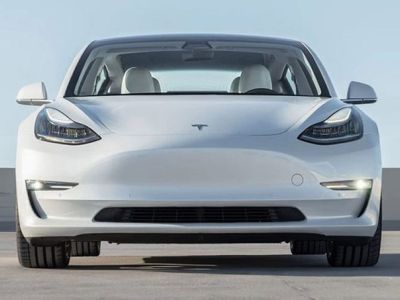 Front view of a white Tesla Model 3 sedan, featuring sleek headlights, a minimalist front design, and a spacious interior, parked on a concrete surface under a clear sky.