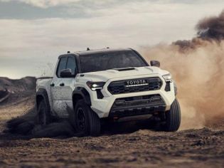 A white Toyota pickup truck drives on a dusty desert terrain, kicking up a cloud of dirt, with rugged hills and a partly cloudy sky in the background.