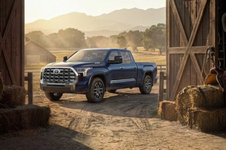 Three-quarter front view of blue Toyota crew cab pickup truck parked on rural farm dirt ground near wooden barn, rolling hills backdrop at golden hour