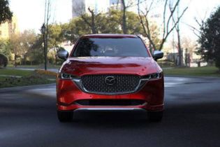 Front view of a red Mazda SUV, featuring its distinctive grille and Mazda logo, parked on a road in a park-like urban setting with trees and buildings in the background.
