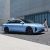 A light blue electric sedan, Xinghai S7, with a sleek design and black wheels, parked near a modern concrete building, with a woman in light clothing walking beside it.