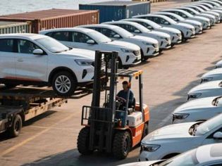 A forklift handling a car at a port with rows of white cars and ships in the background.
