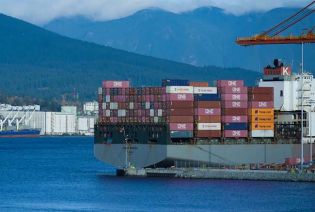 A large container ship loaded with colorful containers docked at a port, with mountains in the background.