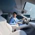 A man in a light blue suit sits in a car's modern interior, featuring light-colored leather seats, a large digital display, and a panoramic roof, highlighting a sleek and tech-forward driving space.