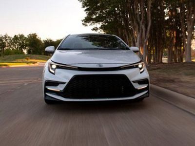 White Toyota sedan driving on road: front view, sleek grille, tree-lined backdrop in soft light.