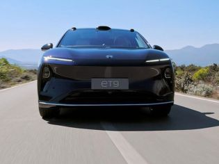 Dark NIO ET9 electric car: front view, driving on road with mountain/vegetation backdrop under blue sky.