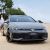 A gray Volkswagen Golf parked on a sandy beach, with a person standing beside it and trees in the background.