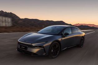 A dark gray Xpeng P7+ electric car driving on a road with mountains and a sunset sky in the background.