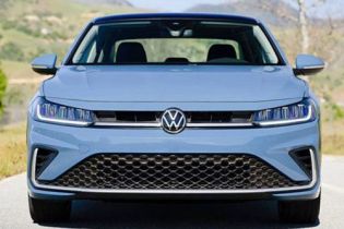 Front view of light blue Volkswagen car: iconic VW logo, sleek LED headlights, grille, on road with green hills.