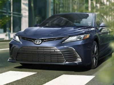 Front view of a dark blue Toyota Camry sedan driving on a city street, with modern buildings and greenery in the background, highlighting its sleek urban design.