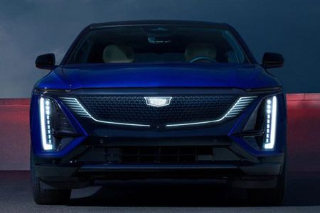 Front view of a blue Cadillac electric vehicle, featuring futuristic LED headlights and a distinctive grille, set against a dramatic nighttime backdrop.