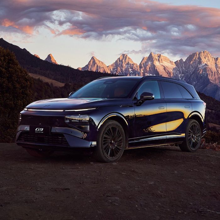 A dark - colored Xpeng G9 SUV parked with a stunning mountain range and a colorful sunset sky in the background.