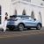 A woman leaning against a light blue SUV parked on the road in front of a white building with arched designs.