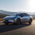 A silver Xpeng P7+ electric car driving on a road with mountains and wind turbines in the background.