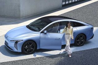 A woman in light clothing exits a light blue electric sedan with a black roof, parked near a modern concrete building, highlighting the car's sleek design and urban sophistication.