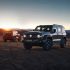 Two off - road SUVs (one gray, one red) parked on an open plain with a sunset and mountainous background.