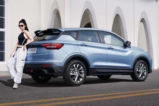 A woman leaning against a light blue SUV parked on the road in front of a white building with arched designs.