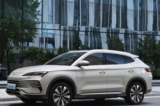 A beige BYD Song PLUS DM-i SUV on an urban road, with a modern glass building and trees in the background, showcasing its sleek design and urban appeal.