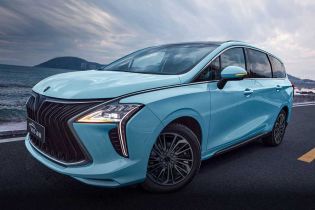 A light blue MPV is parked on a coastal road, with the sea and mountains in the background, showcasing its sleek design and scenic appeal.