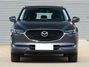 Front view of a dark gray Mazda SUV, parked in front of a corrugated metal wall, featuring its grille, headlights, and sleek front design with a blank license plate.