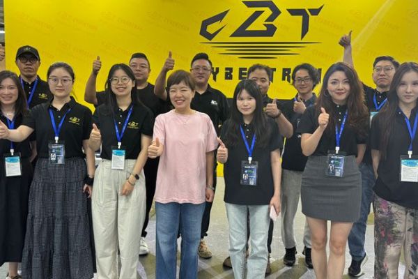 A group of people wearing black "CBT CYBERTRON" shirts, some giving thumbs up, posing in front of a yellow backdrop with the CBT logo, likely at an event.
