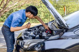 Auto repair technician leaning over a car with its hood open, inspecting and troubleshooting the vehicle engine outdoors in daylight