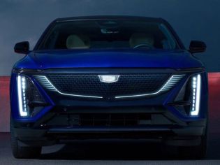 Front view of a blue Cadillac electric vehicle, featuring futuristic LED headlights and a distinctive grille, set against a dramatic nighttime backdrop.