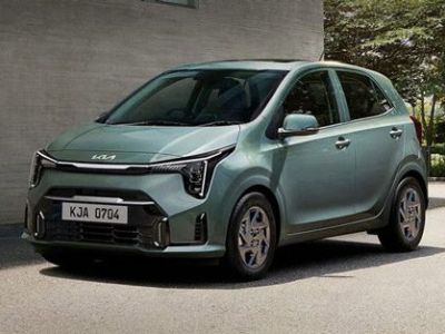A light green Kia hatchback, with license plate KJA 0704, is parked on a paved area beside a concrete building and greenery, showcasing its modern and compact design.