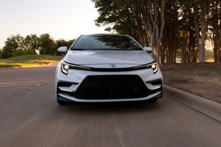 White Toyota sedan driving on road: front view, sleek grille, tree-lined backdrop in soft light.