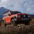 A red off - road SUV parked on a rocky mountain terrain with snow - capped mountains in the background.