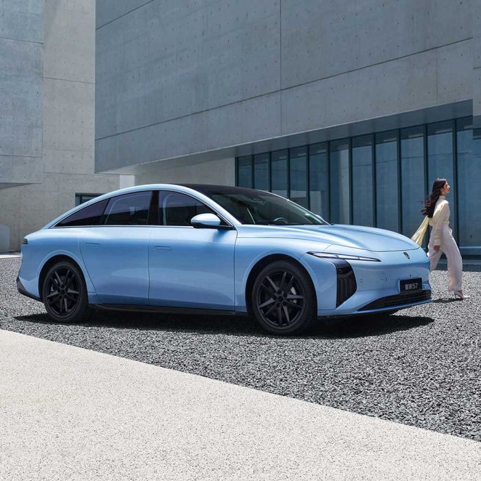 A light blue electric sedan, Xinghai S7, with a sleek design and black wheels, parked near a modern concrete building, with a woman in light clothing walking beside it.