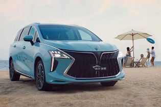 A light blue MPV is parked on a sandy beach, with the sea and a family enjoying the shore in the background, highlighting its stylish design and family-friendly appeal.