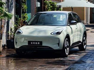 A light green Geely Xingyuan electric car parked on a wet brick road with plants and umbrellas in the background.