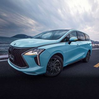A light blue MPV is parked on a coastal road, with the sea and mountains in the background, showcasing its sleek design and scenic appeal.