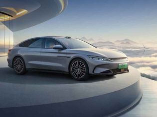 A silver BYD electric sedan displayed on a platform with a futuristic building and cloudscape background.