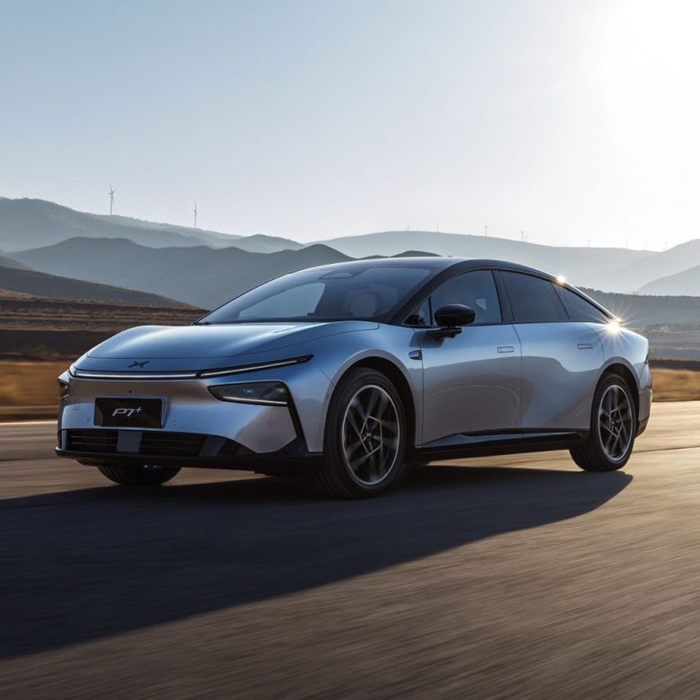 A silver Xpeng P7+ electric car driving on a road with mountains and wind turbines in the background.
