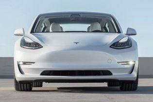 Front view of a white Tesla Model 3 sedan, featuring sleek headlights, a minimalist front design, and a spacious interior, parked on a concrete surface under a clear sky.