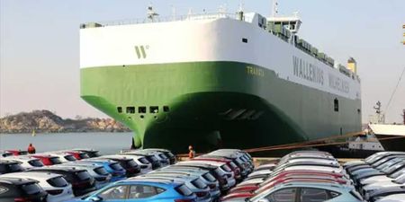 A large green and white Wallenius Wilhelmsen ship docked at a port with many cars parked nearby.