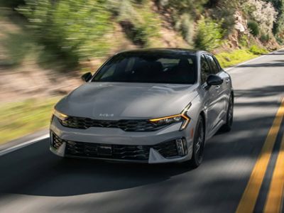 Sporty silver Kia K5 sedan driving on winding mountain road, showcasing aggressive front fascia and signature LED daytime running lights