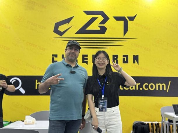 Two people posing in front of a yellow "CBT CYBERTRON" backdrop at an event, one in a teal polo shirt and cap, the other in a black shirt with a lanyard, both making hand gestures.