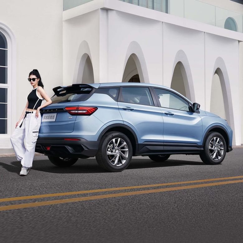 A woman leaning against a light blue SUV parked on the road in front of a white building with arched designs.