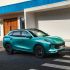 A turquoise Fengxing T5 EVO SUV parked on a crosswalk, with a modern building featuring a blue roof and wooden door in the background, highlighting its sleek side design.