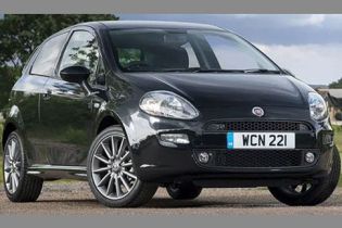 A black Fiat hatchback with license plate "WCN 221", parked on a gravel area with a rural landscape of trees and buildings in the background, showcasing its sleek design.