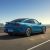 A blue Xiaomi SU7 car driving on a road by the sea under a clear sky.