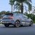 Rear side view of a gray Volkswagen Golf driving on a road, with palm trees and greenery in the background.
